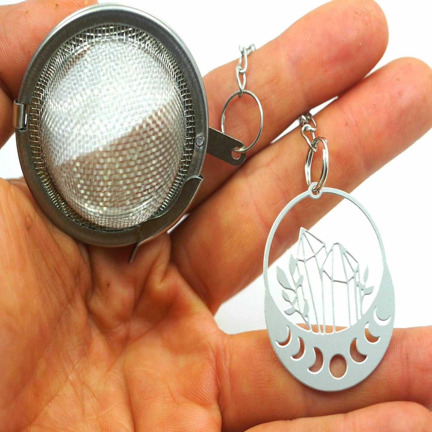 Hand holding a metal tea ball and a crystal-themed keychain against a white background