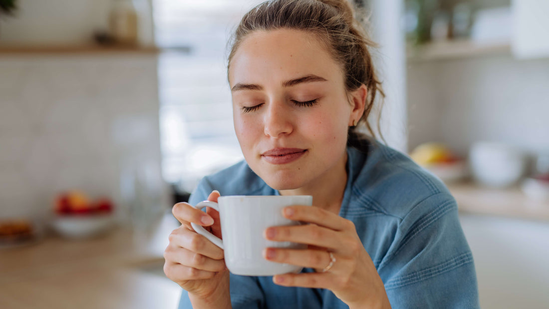 A woman in a blue sweater standing in a kitchen. Her eyes are closed, and she's holding a coffee cup, smiling from the smell or the warmth