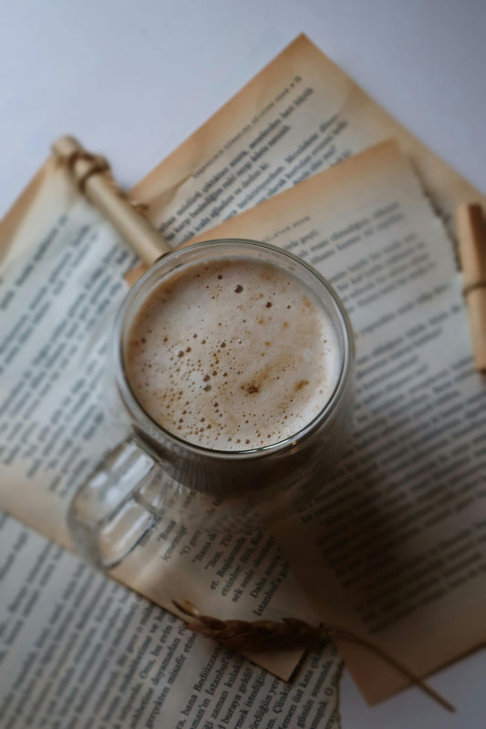 A frothy cup of coffee in a glass mug resting on open book pages, with a warm, cozy aesthetic suggesting a relaxing reading moment.