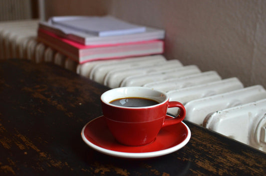 A red mug of coffee sitting on a matching saucer in front of a heater which has books stacked upon it.
