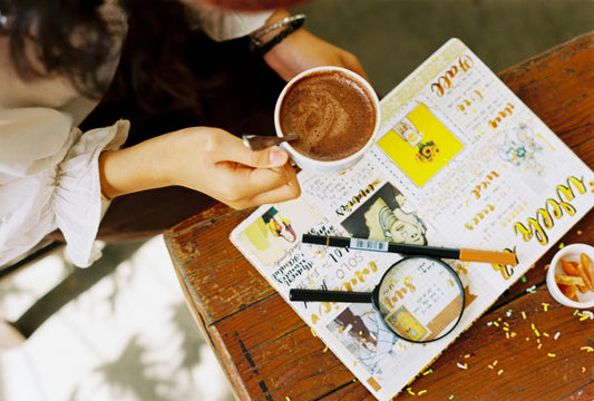 A person holding a cup of coffee while reviewing a creative journal filled with sketches, notes, and colorful designs, with pens and a magnifying glass on a wooden table.