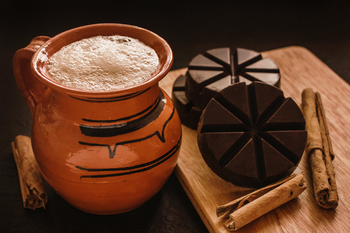 Ceramic pot with frothy liquid, mexican chocolate truffles, and cinnamon sticks on a wooden board.